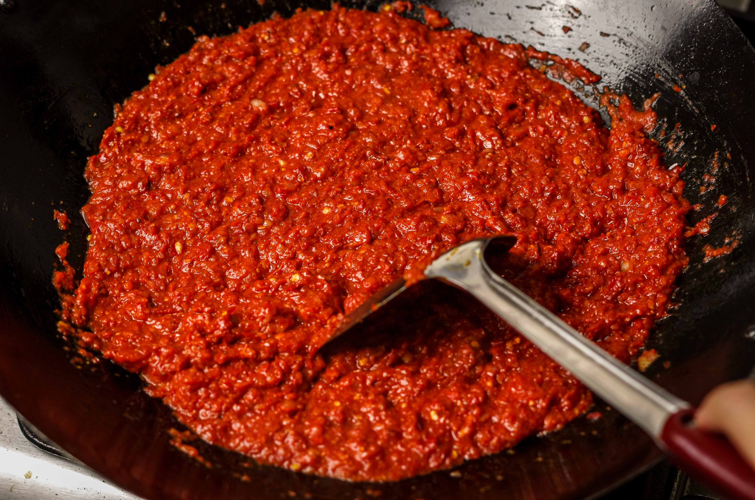 Bright red sambal being stirred with a metal spatula in a black wok.