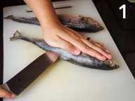A hand is holding down a whole fish on a plastic chopping board. Another hand is holding a knife and making a slit into the fish. A hand is holding down a whole fish on a plastic chopping board. Another hand is holding a knife and making a slit into the fish.