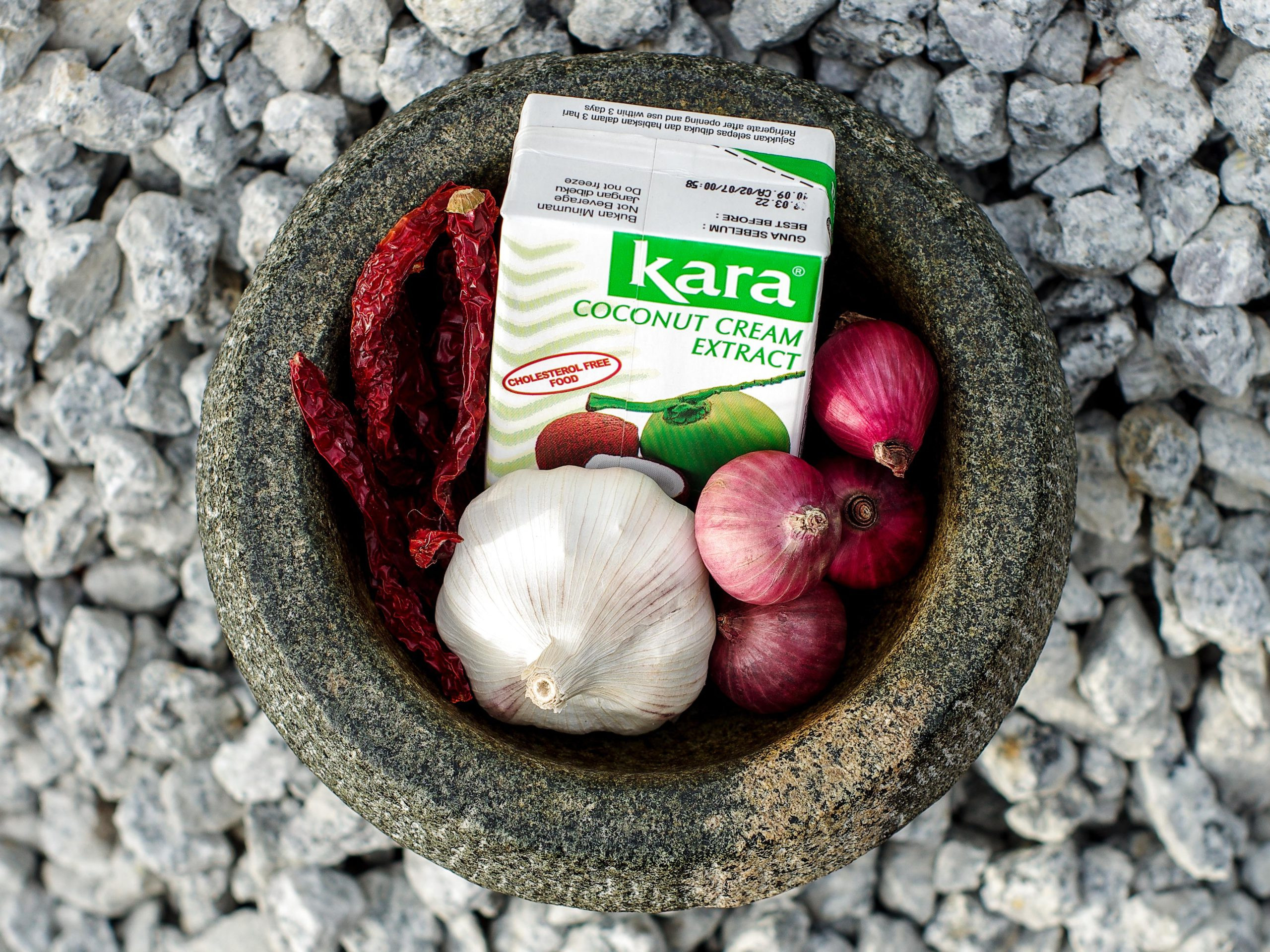A mortar sits on gravel. In the mortar bowl is a box of Kara brand coconut milk, a bulb of garlic, dried chillies, and shallots.