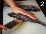 A hand is holding down a whole fish on a plastic chopping board. Another hand is holding a knife and making a slit into the fish. A hand is holding down a whole fish on a plastic chopping board. Another hand is holding a knife and making a slit into the fish.