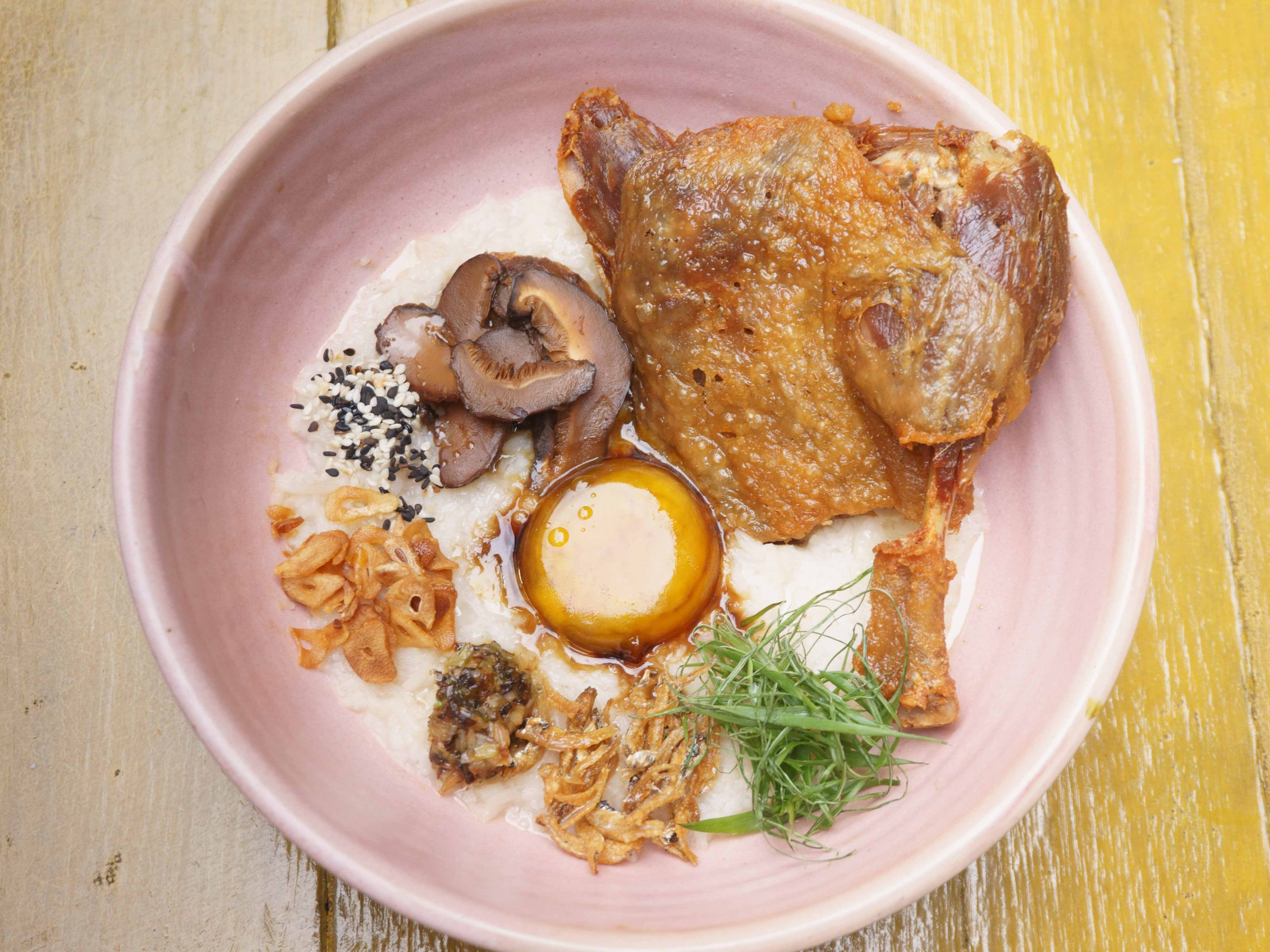 Congee with various toppings including duck confit, in a pink bowl on a wooden table.